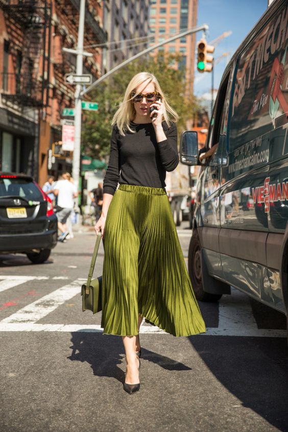 a black top, a green pleated midi skirt, a matching bag and black shoes for a spring work look
