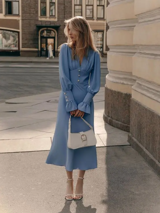 a blue midi dress with a pleated bodice and long sleeves, buttons as details, white shoes and a white baguette bag