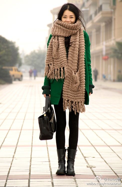 an emerald coat, a beige scarf with two pockets over it and black boots