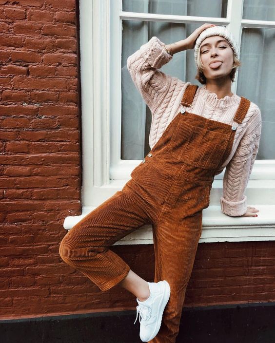 a cable knit neutral sweate and beanie, a rust-colored overall and white sneakers