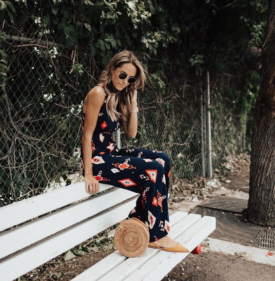 a dark printed jumpsuit with a halter neckline, tan flats and a round wicker bag