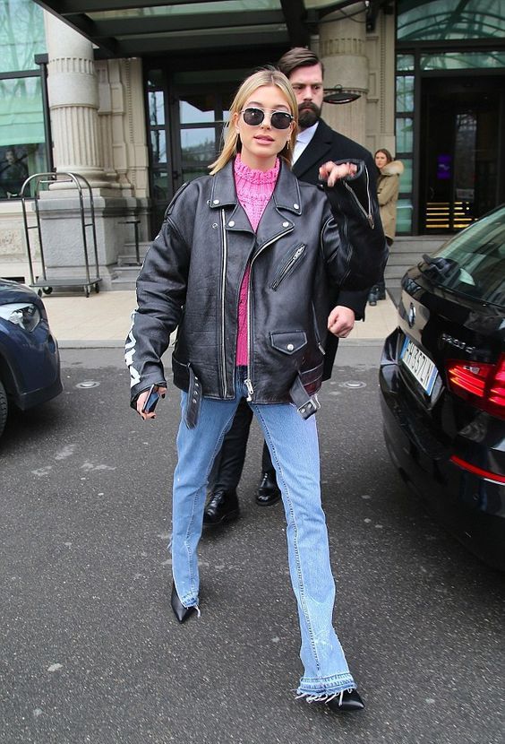blue jeans with a raw hem, black booties, a pink sweater and an oversized leather jacket by Hailey Bieber