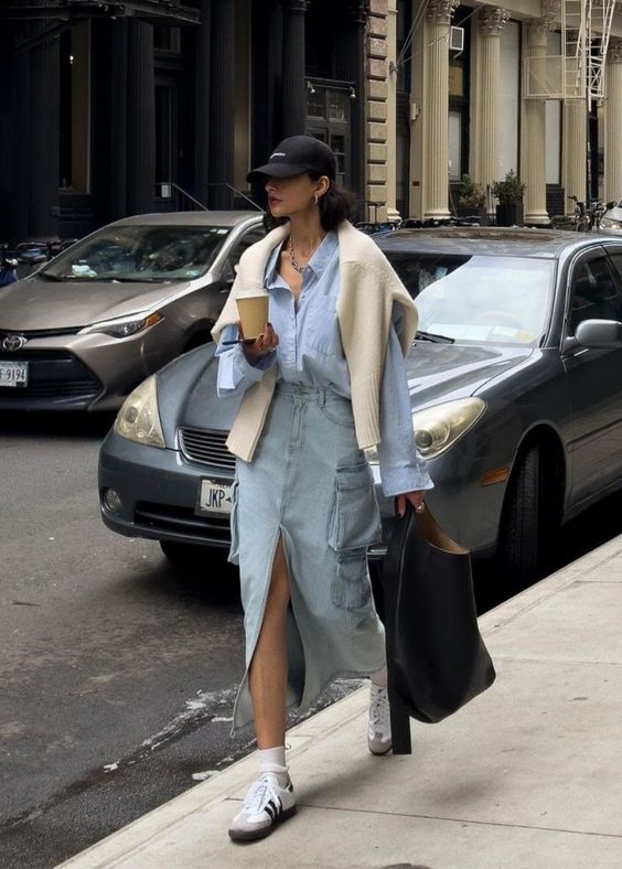 a comfy spring outfit with a blue oversized button down, a blue denim maxi, neutral sneakers and socks, a creamy sweater over the shoulders, a black tote and a black cap