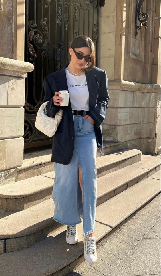 a spring outfit with a printed tee, a bleached denim maxi skirt, a black oversized blazer, white sneakers and a white woven bag