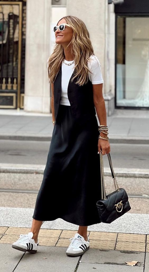 A white t shirt, a black vest, a black satin midi skirt, white sneakers and a black bag with chain
