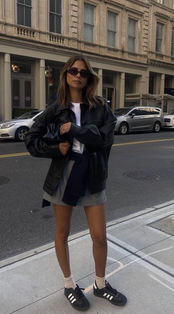 a white t-shirt, a grey mini, a black oversized leather jacket, a black bag and black sneakers plus neutral socks