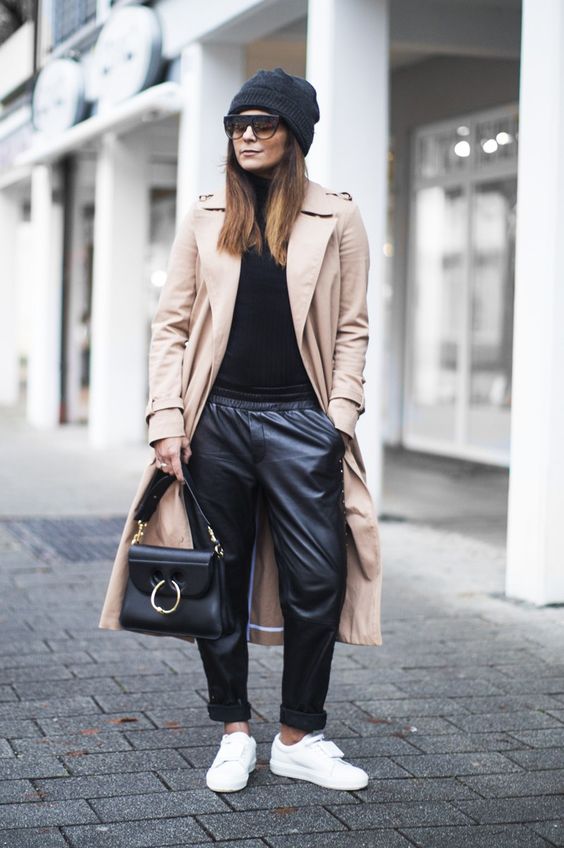 a casual outfit with a black turtleneck, black leather pants, white sneakers, a black bag and beanie and a tan trench