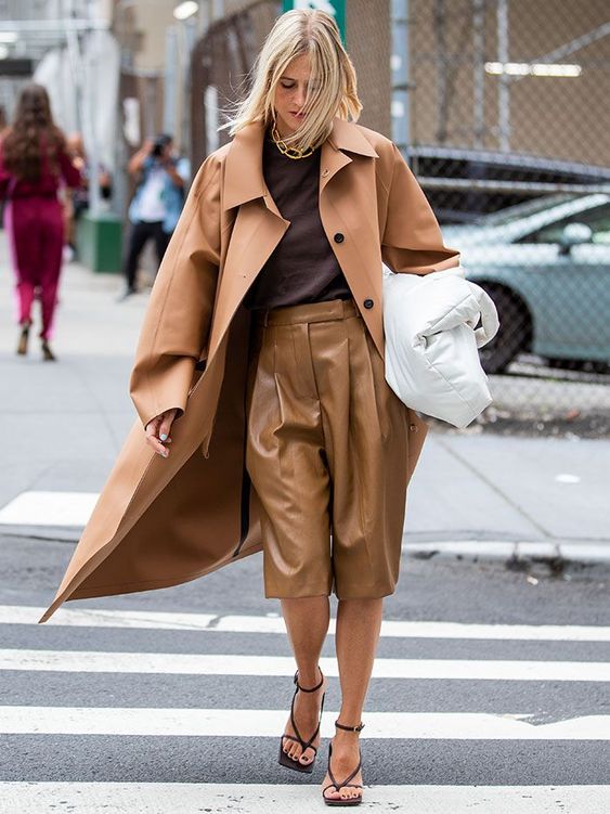 a chic minimalist look with a brown top, tan leather bermudas, black shoes, a brown trench and a white bag