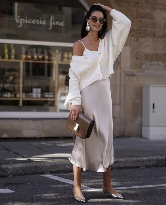 a spring outfit with a white spaghetti strap top, a neutral slip midi skirt, a white cardigan, silver heeled mules and a brown bag