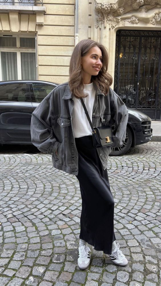 A spring outfit with a white t shirt, a black satin maxi skirt, white sneakers, a grey denim jacket and a black crossbody bag