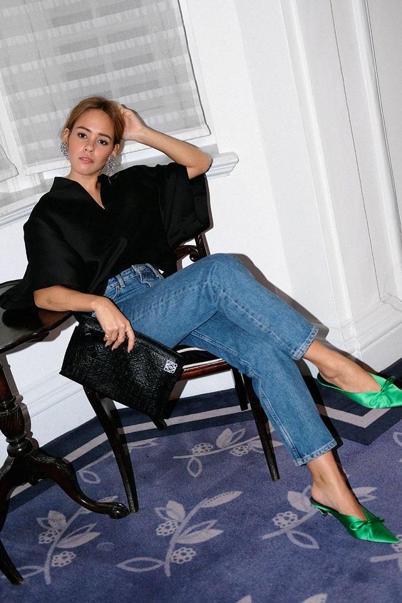 a party outfit with a black blouse with wide sleeves, blue jeans, emerald satin heeled mules and a black bag is wow