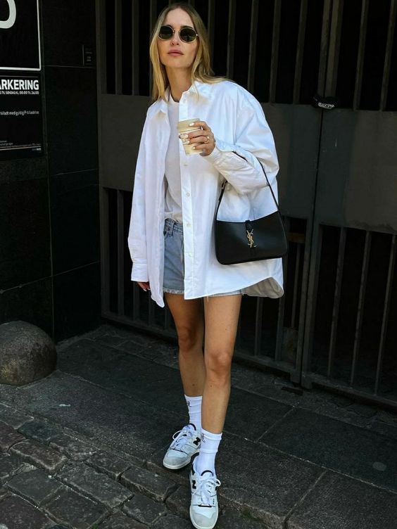 a relaxed summer outfit with blue denim shorts, a white top, an oversized button down, neutral sneakers and socks and a black baguette bag