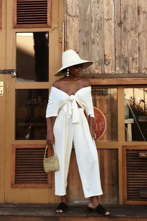 a stylish vacation look with a white off the shoulder tied crop top and matching pants, black slippers and a hat