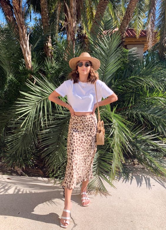 a white cropped tee, a leopard print satin midi, white sandals, a hat and a wicker bag