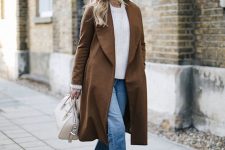 a white oversized sweater, light blue jeans with a raw hem, rust-colored velvet booties and a brown trench