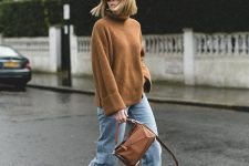 an oversized brown sweater, blue jeans, white ankle booties and a brown geometric bag