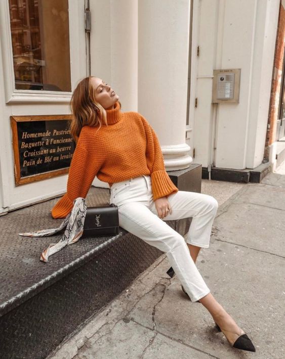 an orange chunky knit sweater with a high collar, white jeans, twon tone shoes and a black bag