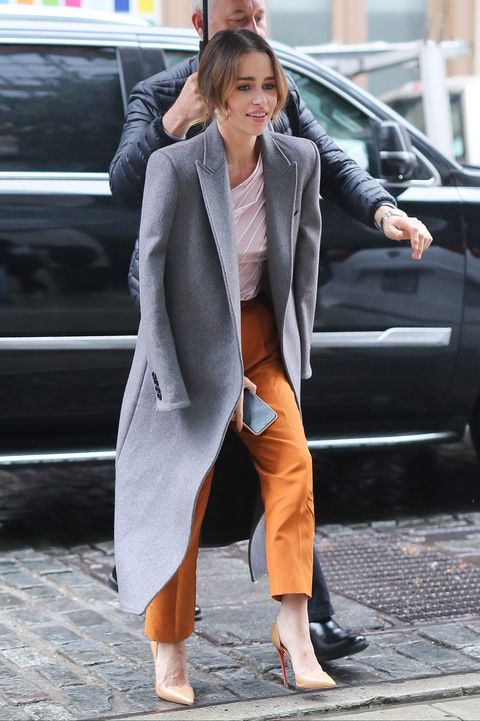 Emilia Clarke wearing a white top, rust-colored pants, tan shoes, a grey midi coat