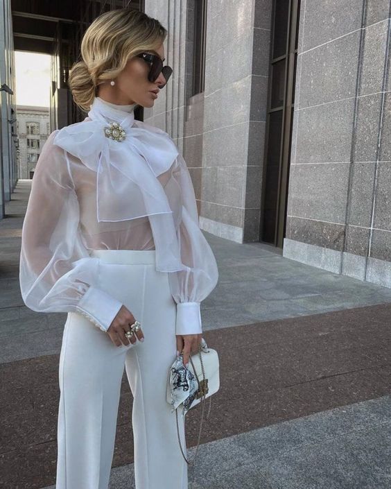 a super elegant look with white pants, a white sheer blouse with a large bow on top, a nude bra and a white bag