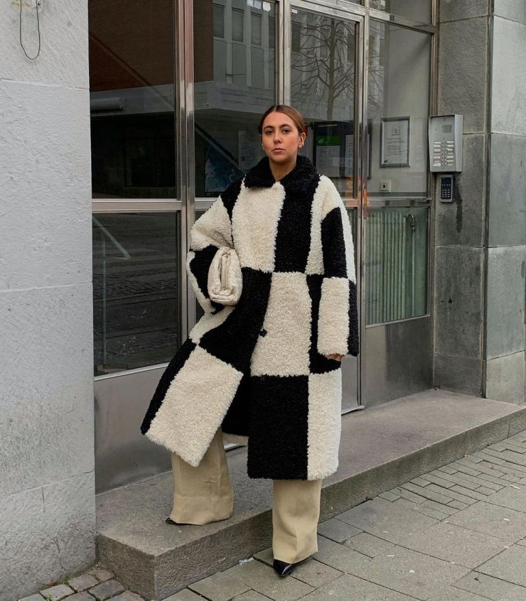 a black and white faux fur coat, neutral pants, black boots and a white oversized clutch