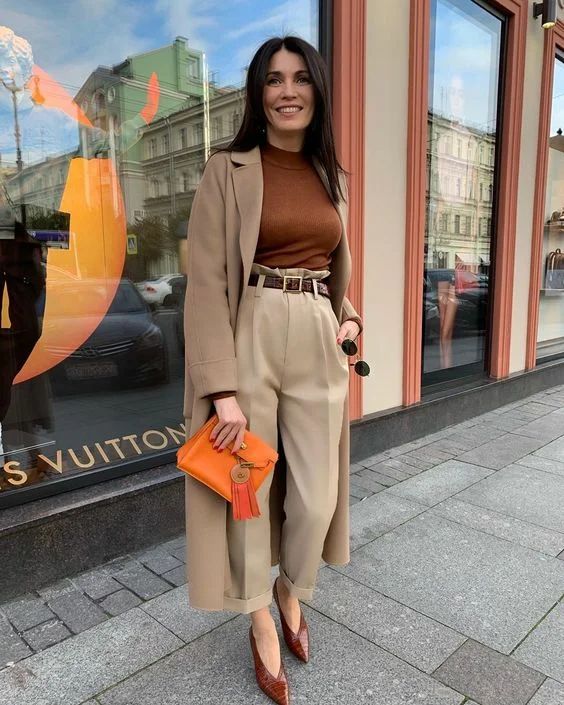 a brown top, tan trousers, a taupe trench, brown vintage-inspired shoes and an orange clutch