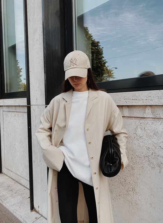 a minimalist look with black pants, a white sweatshirt, a tan trench, a tan baseball hat and a black bag