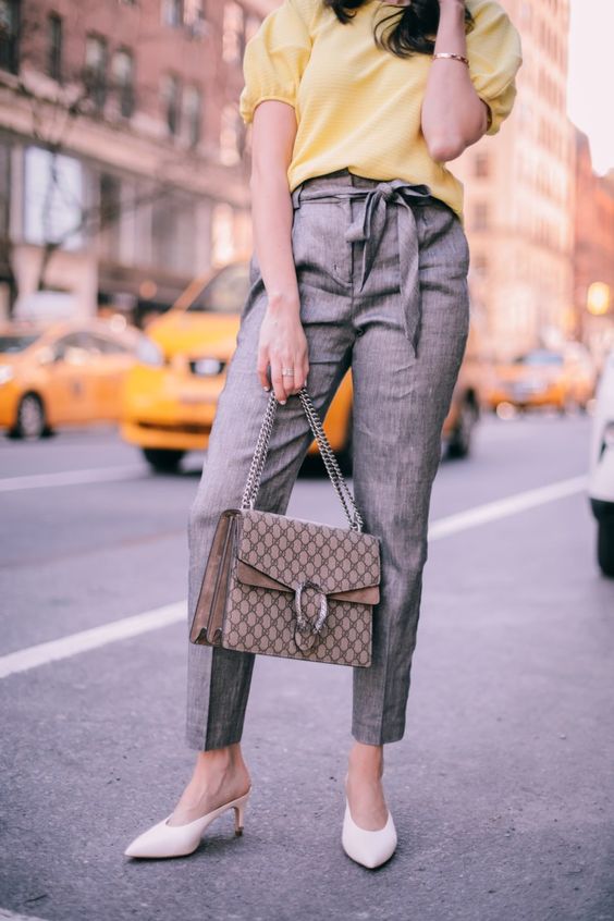 a spring work outfit with a yellow top with puffy sleeves, grey pants, white mules and a printed bag