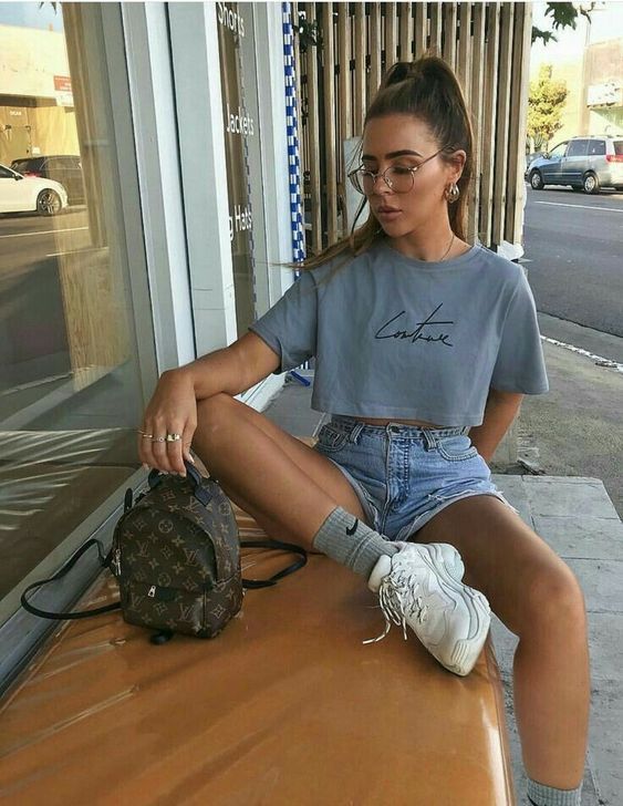 a 90s inspired outfit with a cropped t-shirt, blue denim shorts, white trainers and grey socks plus a brown backpack