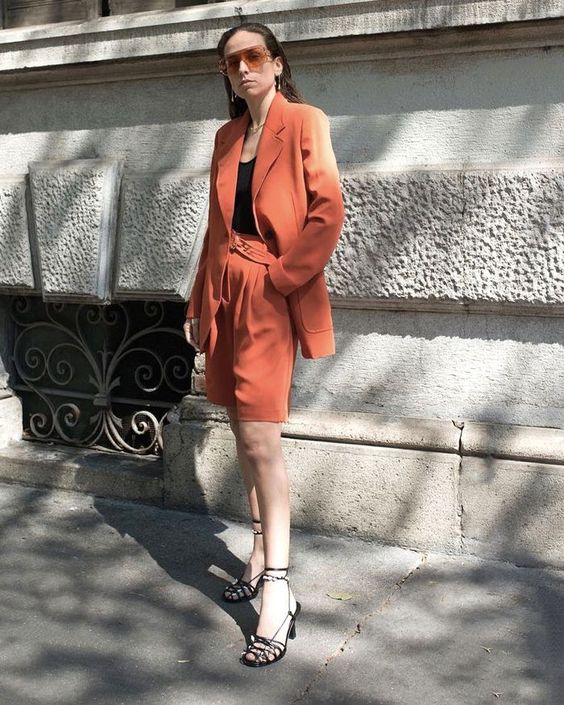 a rust-colored short suit with an oversized blazer and Bermuda shorts, a black top and black lace up heels
