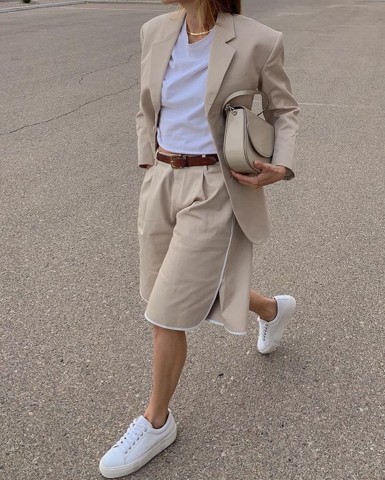 a tan short suit with a fitting blazer and Bermuda shorts, white sneakers and a white t-shirt plus a tan bag