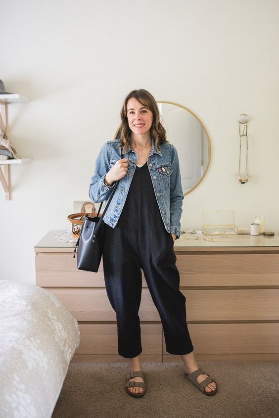 a black jumpsuit, grey birkenstocks, a black bag and a blue denim jacket for a casual work outfit