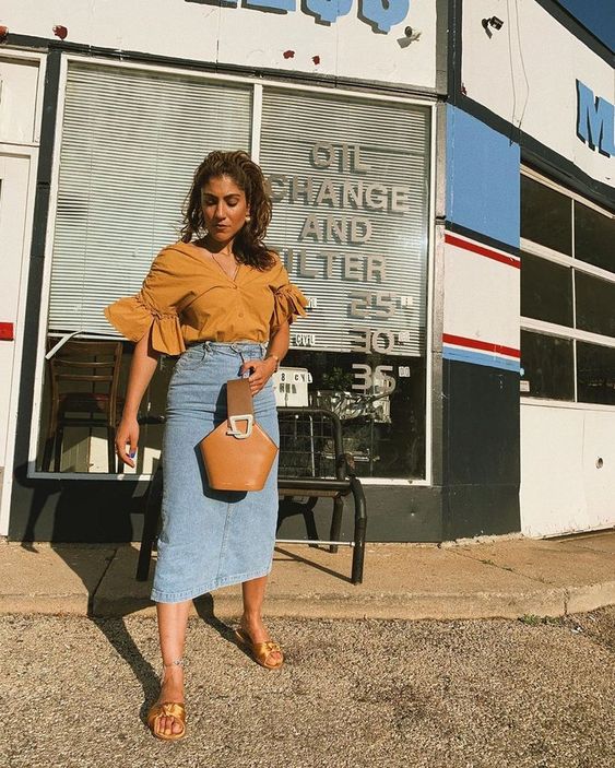 a mustard colored blouse with ruffle sleeves, a blue denim midi, gold mules, a brown bucket bag is amazing
