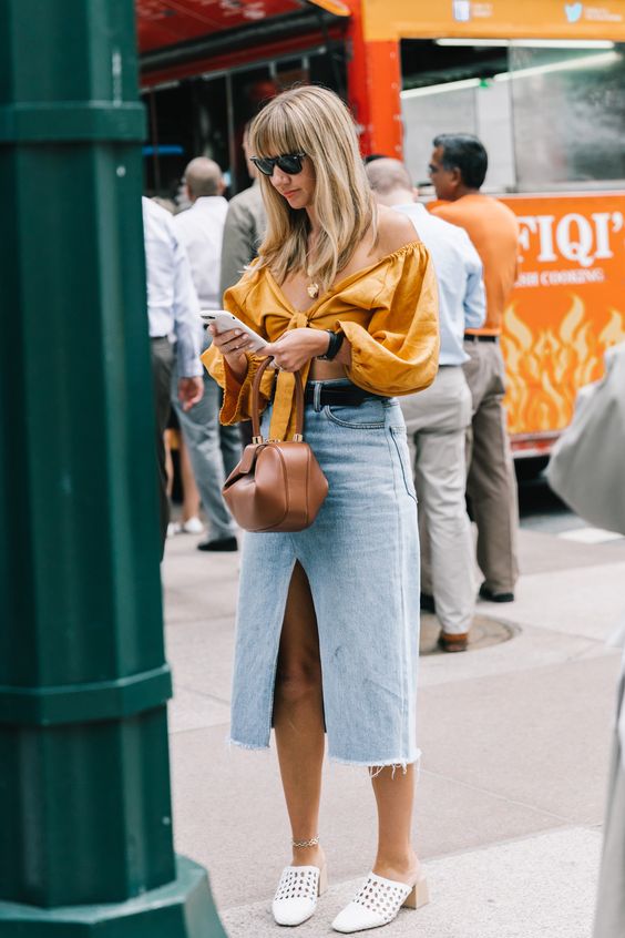 a trendy summer outfit with a mustard off the shoulder crop top, a blue denim midi with a front slit, white mules and a brown bag