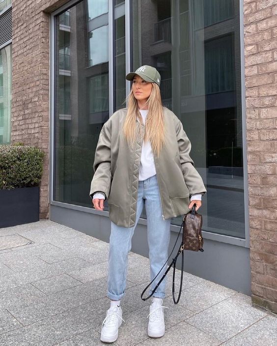 a white sweatshirt, an olive green oversized jacket, blue jeans, white trainers and a green cap