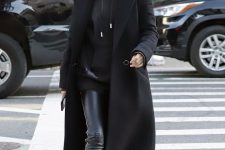 Hailey Bieber wearing a total black look with an oversized hoodie, leather leggings, trainers and a coat plus statement earrings