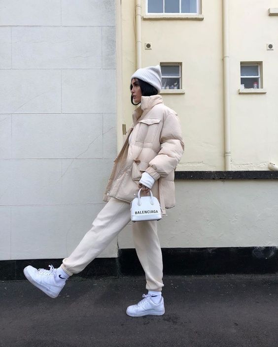 an all neutral sport chic look with creamy sweatpants, a tan puffer jacket, white sneakers, a white beanie and a small bag