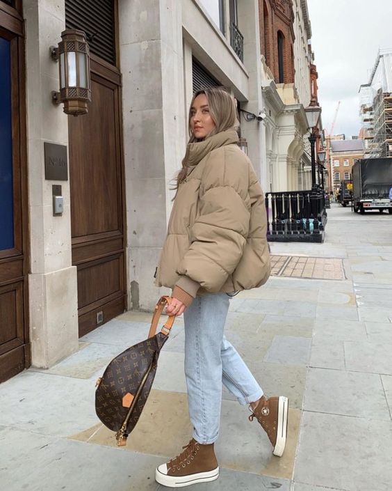 a winter look with a tan puffer jacket, light blue jeans, rust colored sneakers and a brown bag