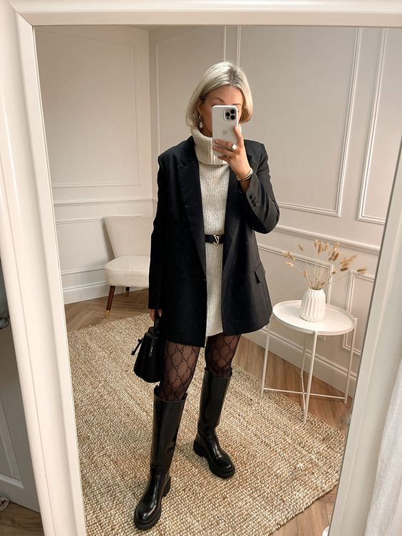 a chic winter outfit with a white sweaterdress, a black blazer and a belt, tights, black riding boots and a bag