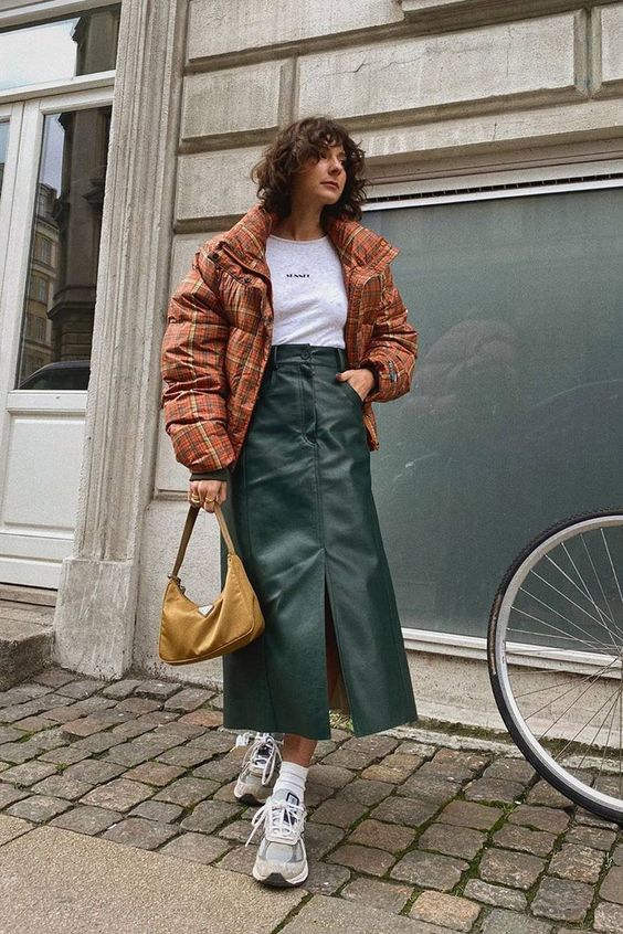 a winter look with a white t shirt, a green A line midi leather skirt with a front slit, trainers, a rust colored puffer jacket and a mustard bag