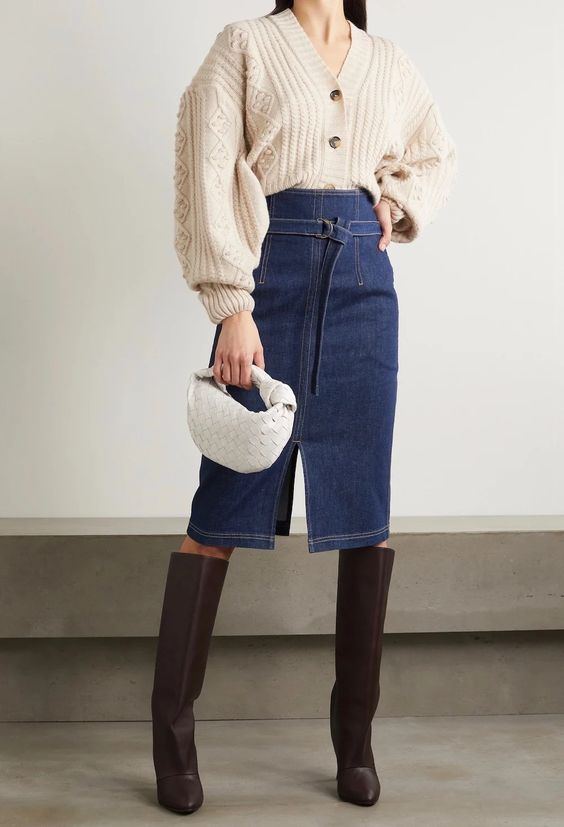 a trendy look with a creamy oversized cardigan as a shirt, a blue high waisted denim midi, brown knee boots and a white woven bag