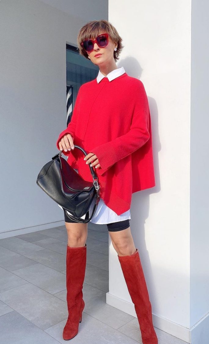 a bold look with leather biker shorts, an oversized white shirt, a colorful asymmetrical red sweater, red knee boots and a black bag