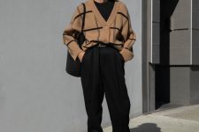 a bold outfit with a black t-shirt, black trousers tucked into black sock boots, a black bag and a beige plaid cardigan tucked inside the pants