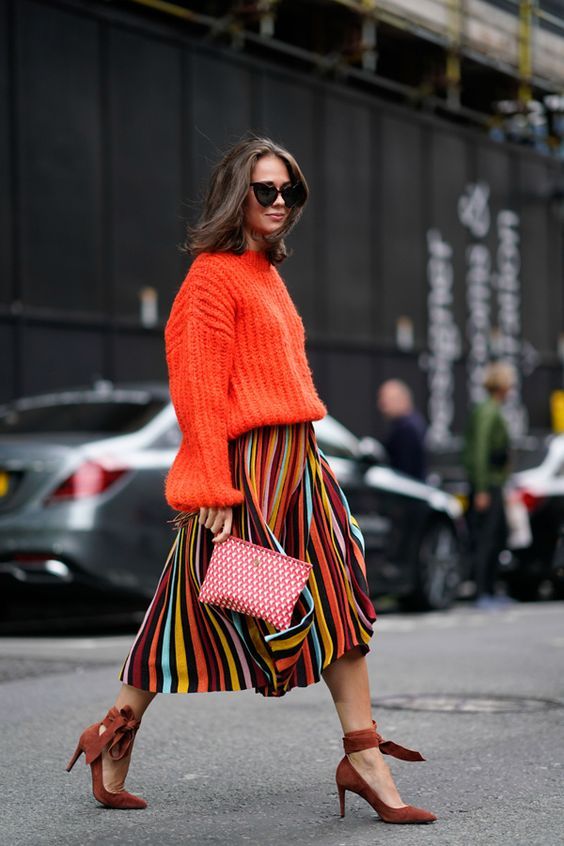 a colorful outfit with an orange chunky sweater and a colorful striped midi skirt, brown sude shoes and a printed clutch