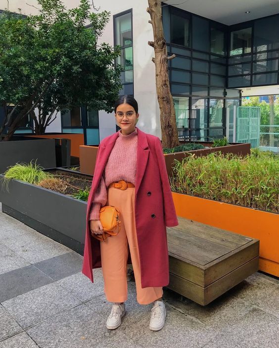 a colorful winter work outfit with a pink ribbed turtleneck sweater, orange trousers, an orange belt and a bag, a fuchsia midi coat