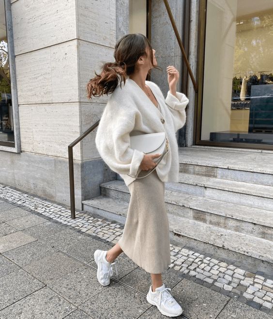 a neutral winter outfit with a white top, a creamy oversized cardigan, a dove grey midi skirt, white trainers and a creamy bag