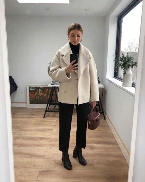 an elegant and minimalist winter work outfit with a black turtleneck, black trousers, boots, a creamy shirt coat and a burgundy bag