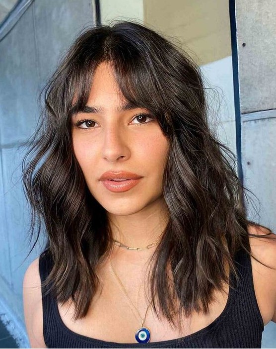 a medium length dark brunette wavy hairstyle with bottleneck bangs and messy waves is a catchy and cool idea to rock right now