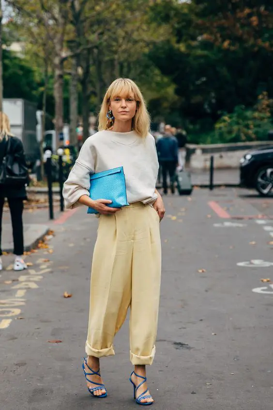 a stylish and delicate look with a touch of color, with a neutral oversized sweatshirt, yellow pleated pants, blue strappy shoes, a blue clutch