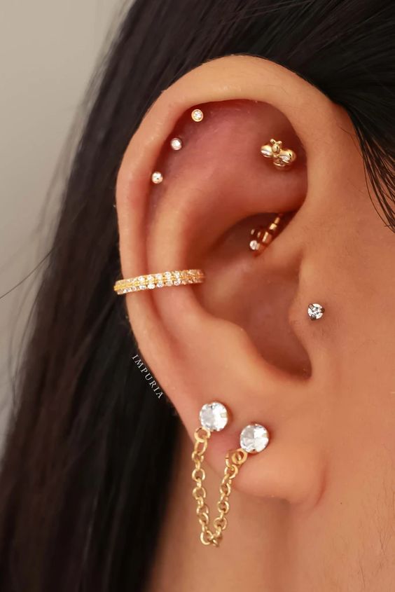 a refined and glam look with a double lobe piercing with chain, a conch piercing with a gold hoop, a tragus and a triple helix piercing with studs and a rook piercing