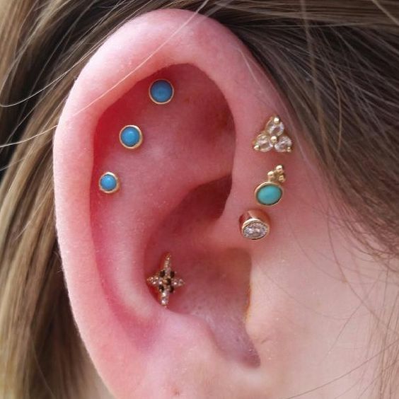 a bold ear with a triple flat piercing done with blue studs, a triple forward helix piercing with mismatching gold studs and a conch piercing